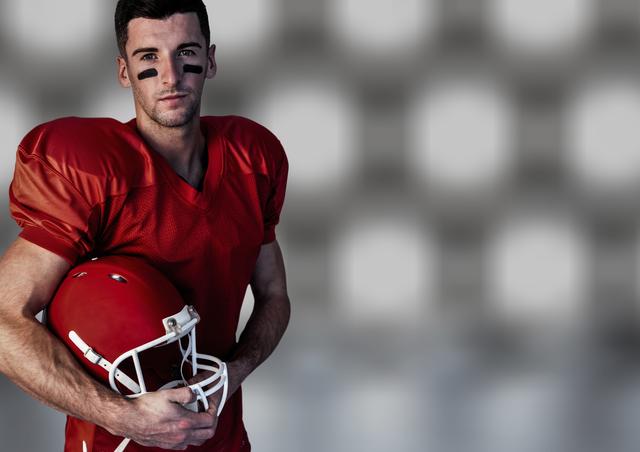 Male American football player in red uniform holding helmet, standing confidently against a blurred background. Ideal for sports promotions, athletic advertisements, and motivational posters.