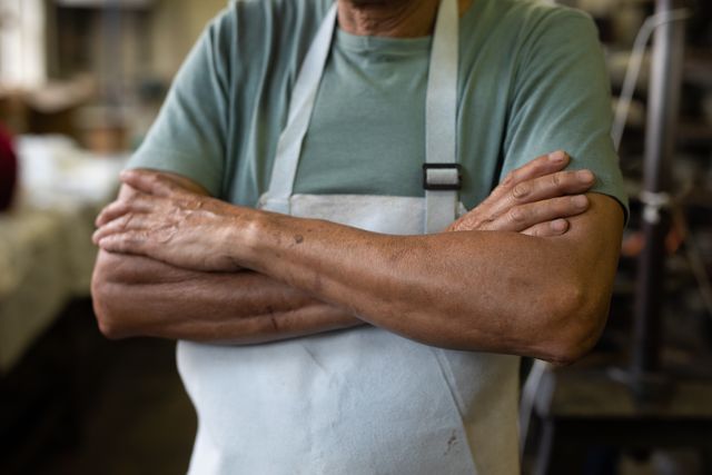 Mid section close up of a senior biracial man with crossed arms standing in the workshop at a hat factory wearing an apron.
