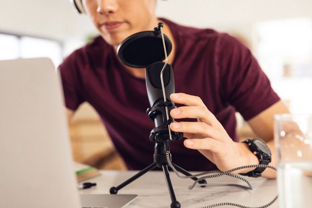 Teenage boy using a microphone and laptop at home, suggesting a modern lifestyle involving technology. Ideal for content related to podcasting, remote work, online communication, digital content creation, and home-based activities.