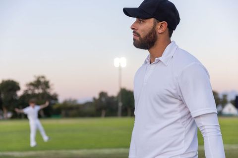 Bearded cricketer standing at dusk under floodlights wearing white kit and black cap