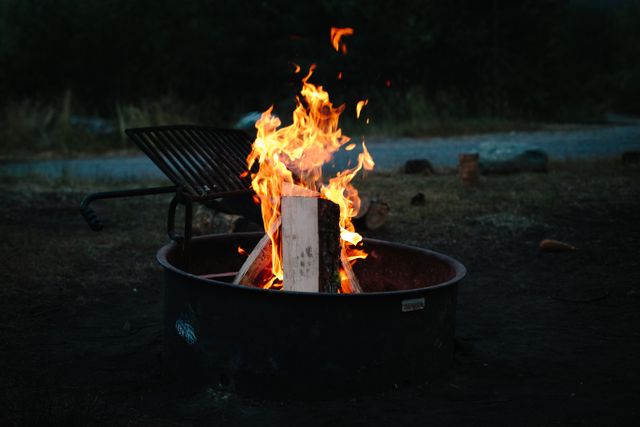 Bright fire burning wood in metal fire pit at campsite. Ideal for depicting outdoor camping experiences, family gatherings, or nature retreats. Flames create a warm, inviting atmosphere perfect for travel blogs, camping gear advertisements, or outdoor event promotions.