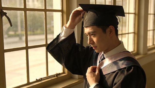 Asian graduate adjusting mortarboard and sash by sunlit campus window, thoughtful portrait