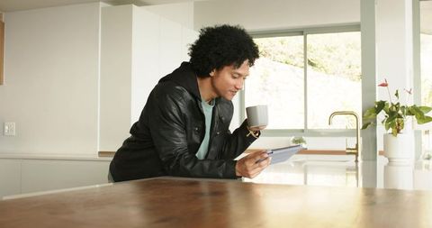 African American man leaning on kitchen counter using tablet and holding coffee mug