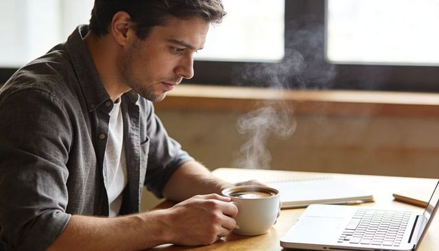 Focused man working on laptop while holding steaming coffee cup at window-side wooden desk