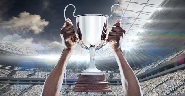 Hands of an athlete holding a championship trophy in a large stadium filled with spectators. Use for sports victory celebrations, achievements, motivation, or promotional material for athletic events.