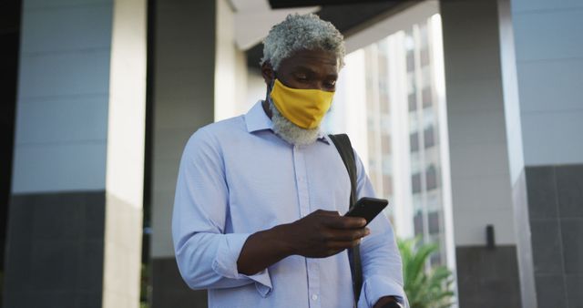 African american senior man wearing face mask using smartphone and looking around in corporate park. hygiene and social distancing during coronavirus covid-19 pandemic.