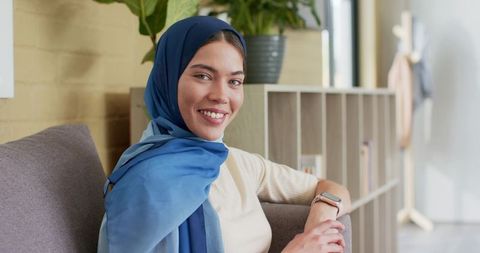 Smiling indian woman wearing blue hijab relaxing on sofa in modern living room