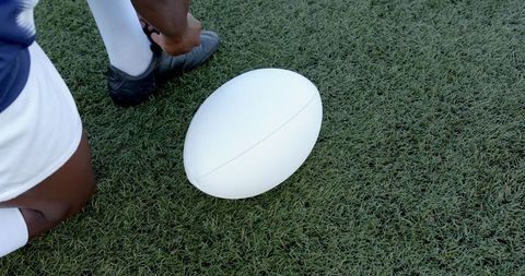 Rugby player tying cleat beside white ball on turf field