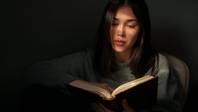 Woman reading hardcover book by warm lamplight in moody chiaroscuro interior