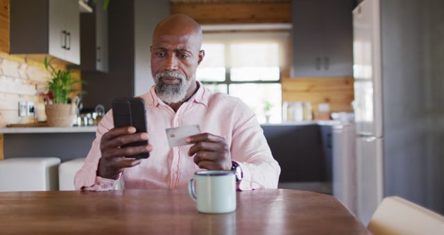 Senior man using smartphone and credit card while sitting at table in modern kitchen, demonstrating everyday online shopping routine. Ideal for illustrating concepts of e-commerce, online payments, and digital transactions targeting senior market. Could be used in advertisements, promotional content, or articles about technology adoption among older adults.