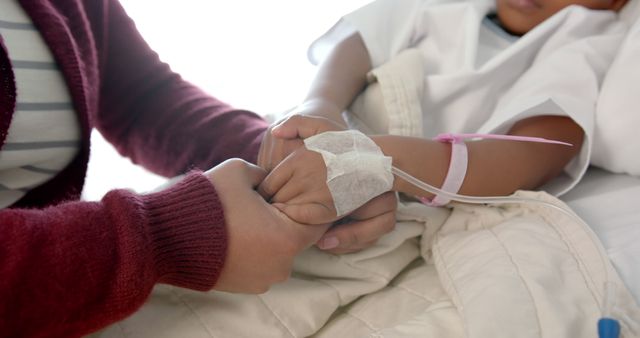 Midsection of african american girl patient and her mother holding hands in bed in hospital room. Medicine, healthcare and hospital.