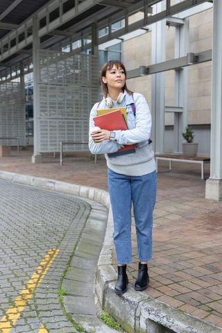 Asian female student standing on campus curb holding notebooks wearing headphones