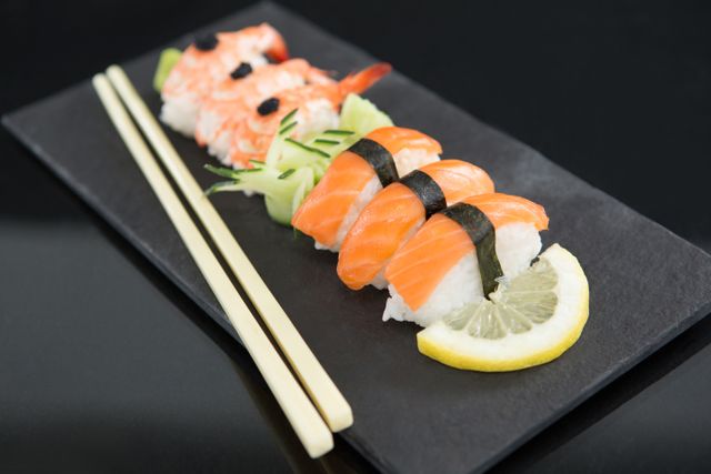 Close-up featuring a sushi platter with nigiri and shrimp roll on a black slate plate, accompanied by chopsticks and a lemon slice garnish. Ideal for use in culinary blogs, restaurant menus, or food advertisements showcasing Japanese cuisine.