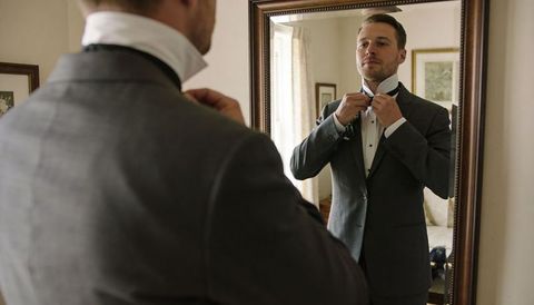 Adjusting bow tie in mirror preparing groom in dark suit and boutonniere for wedding