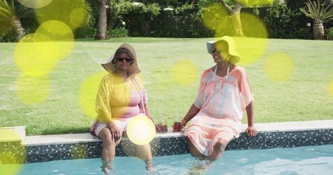 Mature women relaxing poolside wearing sun hats and summer dresses sharing cold drinks