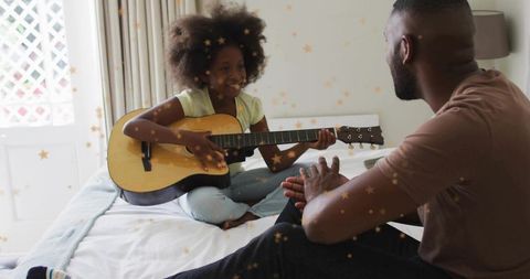 Young girl playing acoustic guitar on bed while father clapping in warm music moment