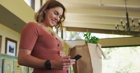 Pregnant Woman Checking Smartphone with Grocery Bag in Kitchen