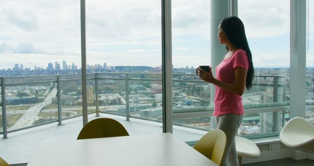 Woman stands in bright office with large windows, holding coffee and looking at distant city skyline. Ideal for themes of work-life balance, modern architecture, urban living, mindfulness, professional life, office interiors.