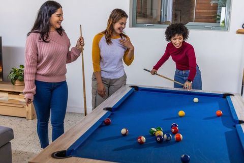 Diverse women playing billiards and laughing around blue-felt pool table at home