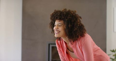 Smiling woman leaning forward in cozy living room wearing textured pink top, curly hair, fireplace