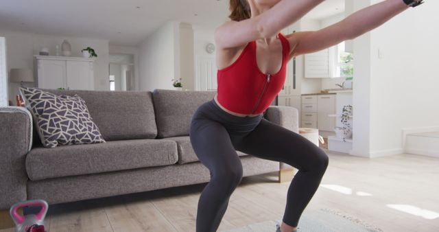 Caucasian woman wearing red sports suit and doing squats at home. Domestic life, healthy lifestyle and fitness, unaltered.