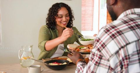Woman serving salad to partner at sunlit wooden table, couple sharing cozy meal