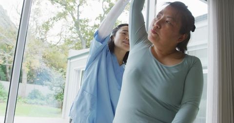 Caregiver assisting woman with arm stretch during home therapy by sliding glass door