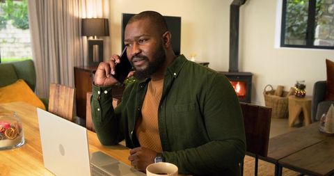 Mid-30s African American man talking on phone while working on laptop in cozy home office with firep