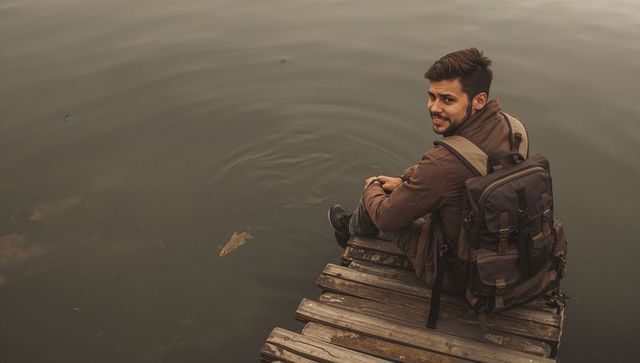 Traveler sitting on weathered wooden jetty looking back over moody lake with backpack