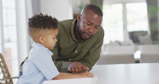 Happy african american man and his son sitting at table and reading braille. Spending quality time at home, childhood, blindness and family concept.