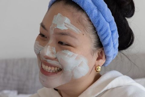 Asian woman smiling wearing creamy facial mask and blue headband at-home skincare routine