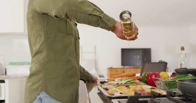 This image portrays a man in a casual green shirt pouring olive oil into a roasting tray filled with fresh vegetables in a modern kitchen. The focus on the use of fresh ingredients and healthy cooking makes it perfect for blogs, articles, and advertisements related to healthy lifestyles, culinary arts, and cooking tutorials. It is also useful for illustrating themes of domestic life and modern home interiors.