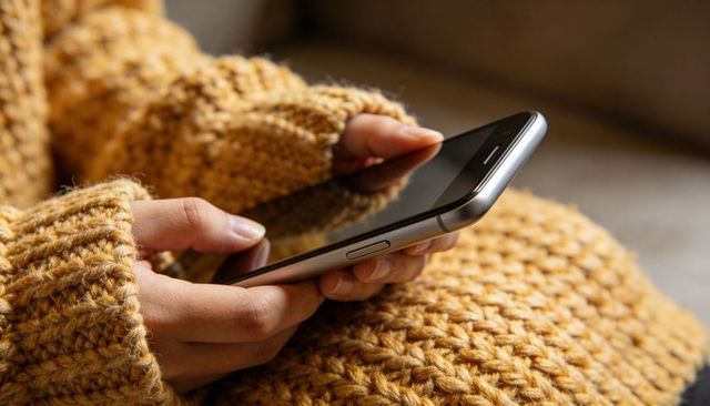 Holding smartphone on lap with mustard chunky knit sweater, cozy hands scrolling