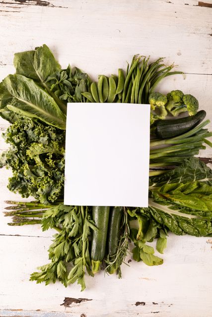 Blank white paper placed on a variety of fresh green leafy vegetables including kale, lettuce, and zucchini. Ideal for use in healthy eating promotions, organic food advertisements, diet and nutrition blogs, and wellness campaigns. The blank space can be used for adding text or logos for marketing purposes.