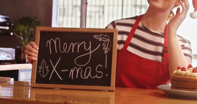 Happy barista displaying cheerful Merry Christmas message on chalkboard in cozy café. Perfect for holiday promotions, festive greetings, cafe advertisements, and spreading holiday cheer.