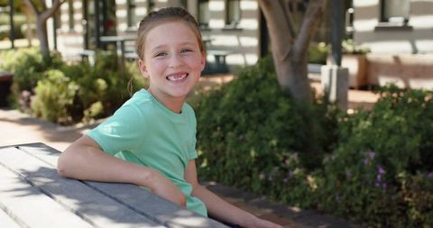 Cheerful elementary girl smiling while sitting on picnic bench in sunny courtyard