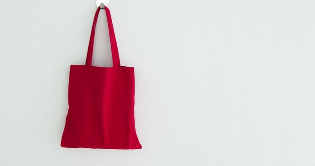 A vibrant red tote bag hangs on a white wall, with copy space. Its bold color stands out, suggesting a focus on eco-friendly fashion or reusable shopping bags.