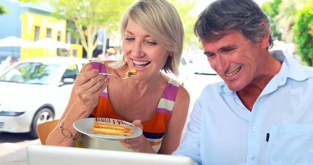 File depicts a joyful senior couple enjoying dessert while browsing on a laptop at an outdoor café. Can be used in content promoting senior lifestyle, retirement activities, leisure activities, or healthy aging. Ideal for articles or ads revolving around happiness, technology use among seniors, or casual dining experiences.