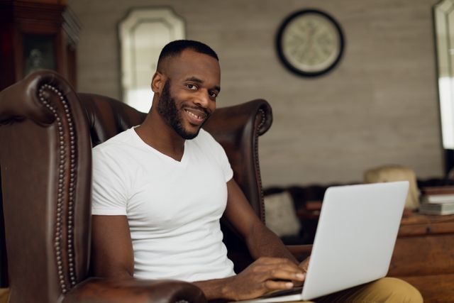 Side view of an African-American smiling and sitting on a leather chair inside a room while using a laptop