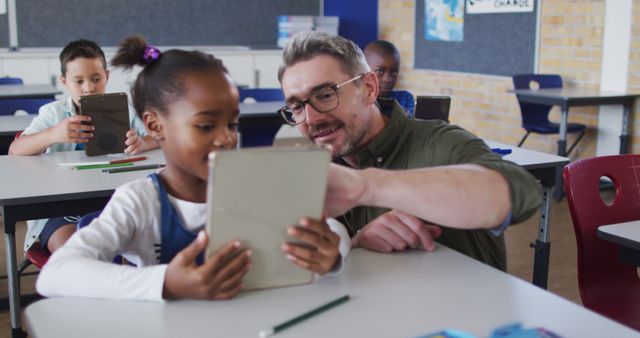 Shows a teacher assisting a young student using a tablet in a diverse classroom setting. Ideal for educational resources, technology in education, teacher training, digital learning materials, and school advertisements.