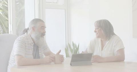 Mature couple chatting over coffee and tablet at sunlit table, relaxed conversation