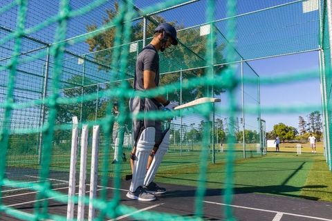 Young cricketer practicing at bat in netted training cage on sunny outfield with teammates