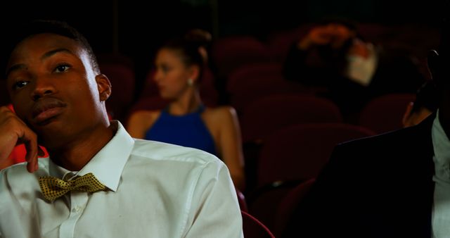 A young African American man appears bored or disinterested while sitting in a theater, with copy space. His expression contrasts with the other audience members who are engaged or reacting differently to the performance or movie.