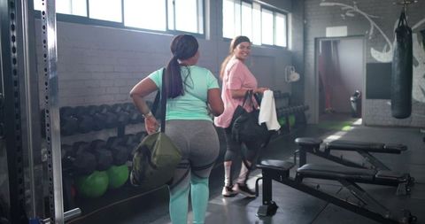 Diverse women walking through gym weight area carrying duffel bags, smiling and confident