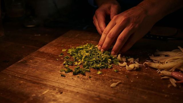 Adult male hands pressing chopped chives on worn wooden board with onions in warm light