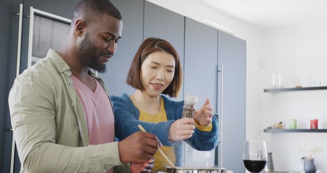 Multicultural couple preparing meal in modern kitchen. Woman uses pepper grinder while man stirs ingredients in pot. A glass of red wine is on the counter. Ideal for lifestyle, culinary blogs, home cooking guides, and promoting family or romantic cooking experiences.