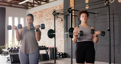 Two women lifting hex dumbbells during partner strength training in bright industrial gym