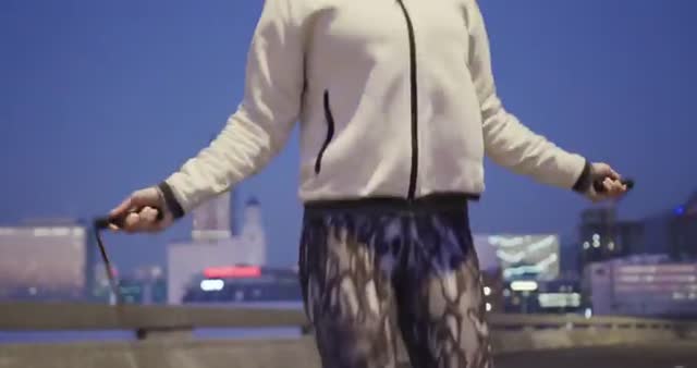 This image depicts a fit Caucasian woman engaging in a night-time workout by skipping rope amidst a city backdrop. The focus on her athletic attire and activity makes it ideal for websites and promotional materials related to fitness, exercise routines, urban lifestyle, or sports apparel. It can be used to inspire an active lifestyle or as an illustrative visual for sports-related articles and blogs.