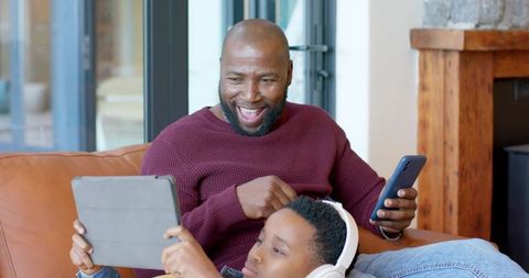 African american father and son sharing tablet, smiling and relaxing on cozy leather couch