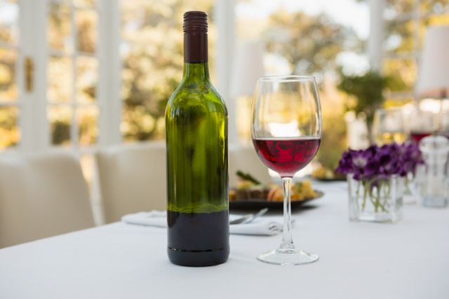 Close up of a wineglass filled with red wine and a green wine bottle on a white tablecloth in a restaurant. Ideal for use in advertisements for restaurants, wine brands, dining experiences, or romantic dinner settings. Perfect for illustrating concepts of luxury, elegance, and fine dining.